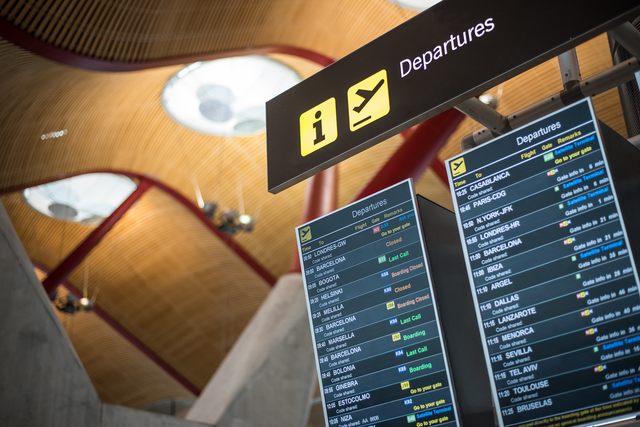 Electronic departure board glowing above a crowded airport terminal, listing flights and delays as travellers queue below.